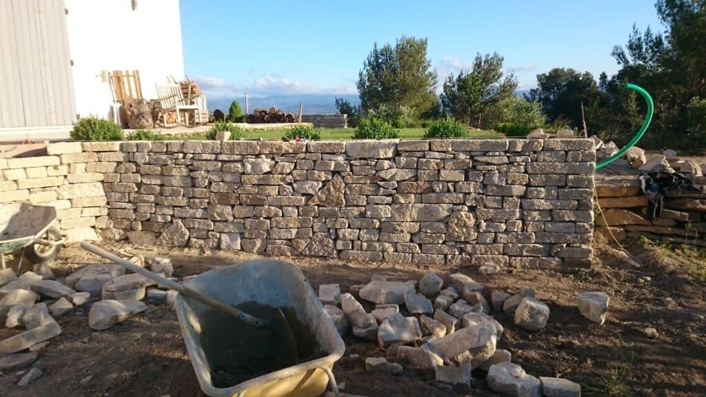 Construction d'un Mur en Pierres Sèches pour le Jardin Construction d'un mur en pierre naturelle avec brouette de mortier et pierres de taille dans un jardin ensoleillé.