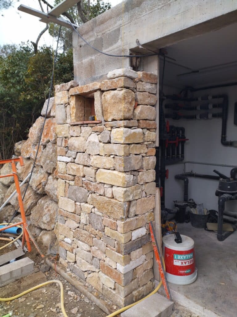 Construction d'un mur en pierre sèche avec niche, fils à plomb et seau Relax Malaxe devant un local technique ouvert.