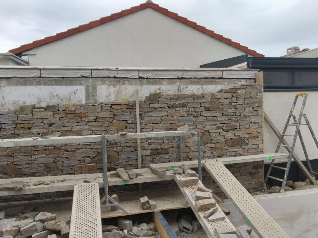 Construction d'un mur en pierre naturelle sur chantier Mur en pierre naturelle en construction avec échafaudages et échelle devant une façade de maison.