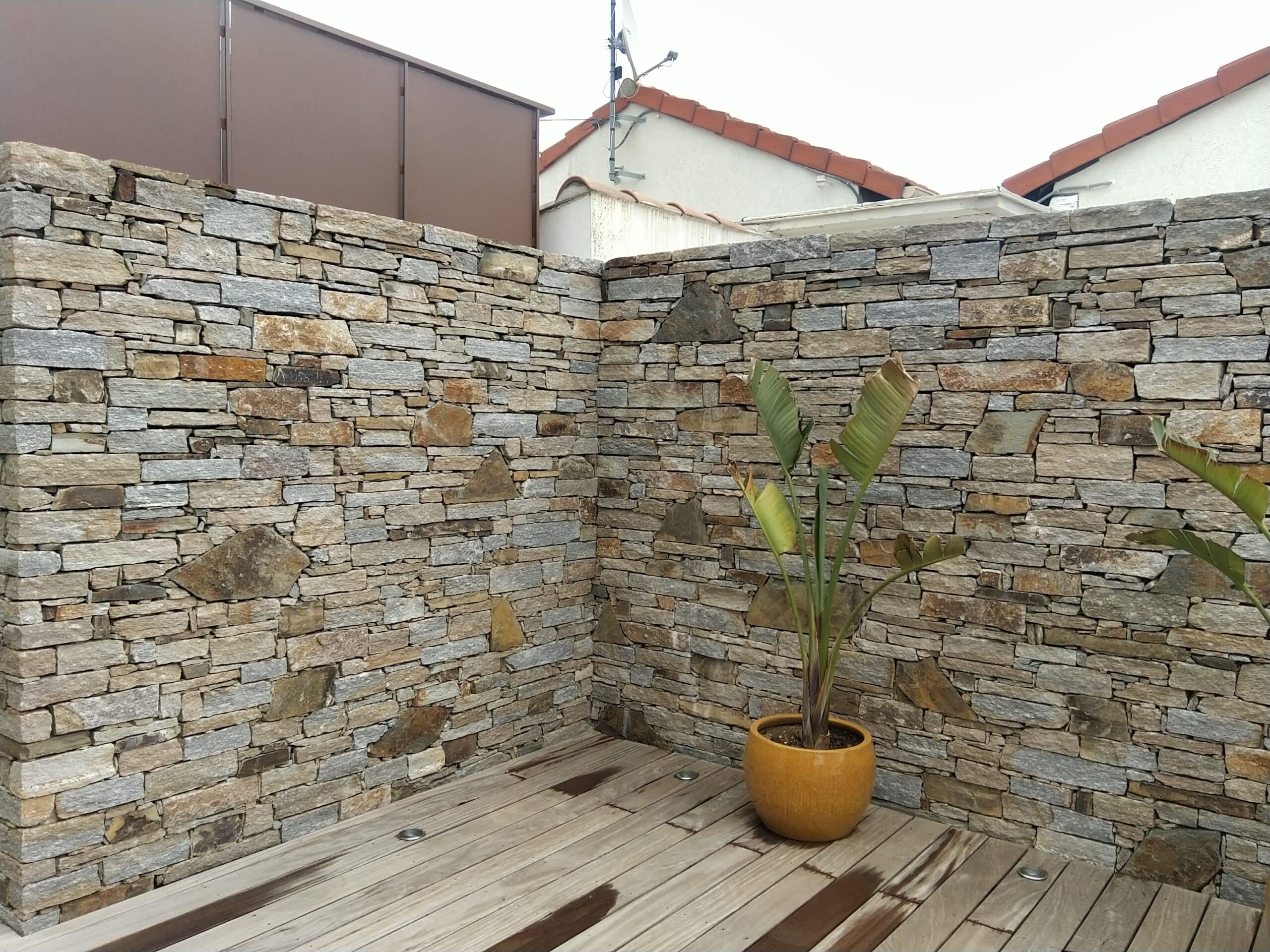 Mur en Pierre Naturelle et Aménagement Terrasse Bois Mur de parement en pierre naturelle et terrasse bois avec plante exotique Strelitzia en pot jaune.