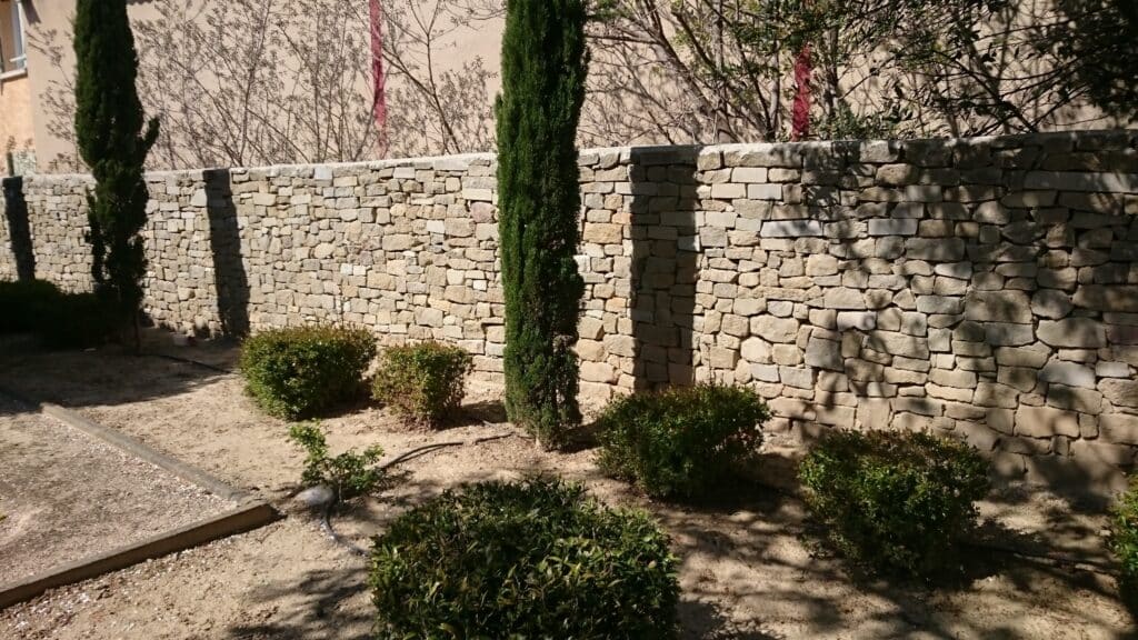 Mur en pierres sèches avec cyprès et buissons verts dans un jardin méditerranéen ensoleillé.