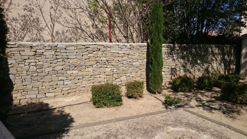 Mur en pierre sèche dans un jardin ensoleillé avec un cyprès élancé et des arbustes verts.