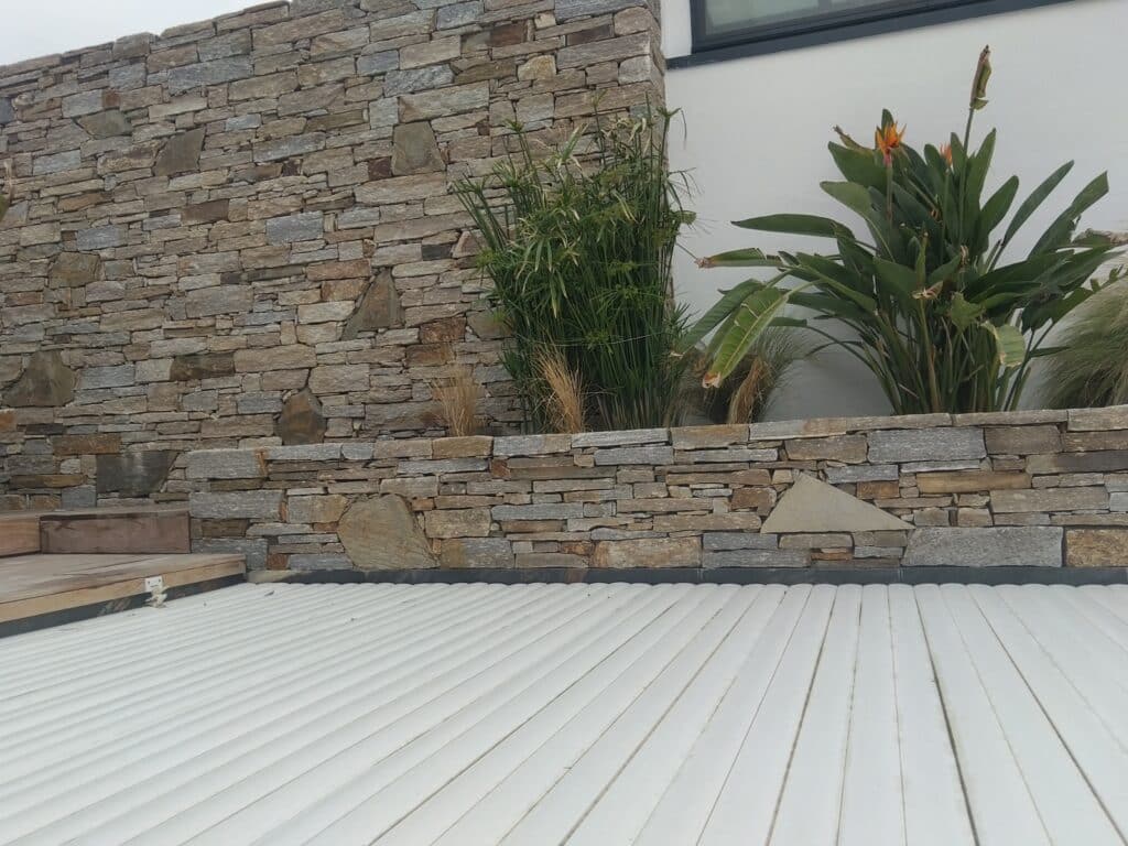 Mur en pierre sèche bordant une piscine avec volet roulant blanc et plante oiseau du paradis.