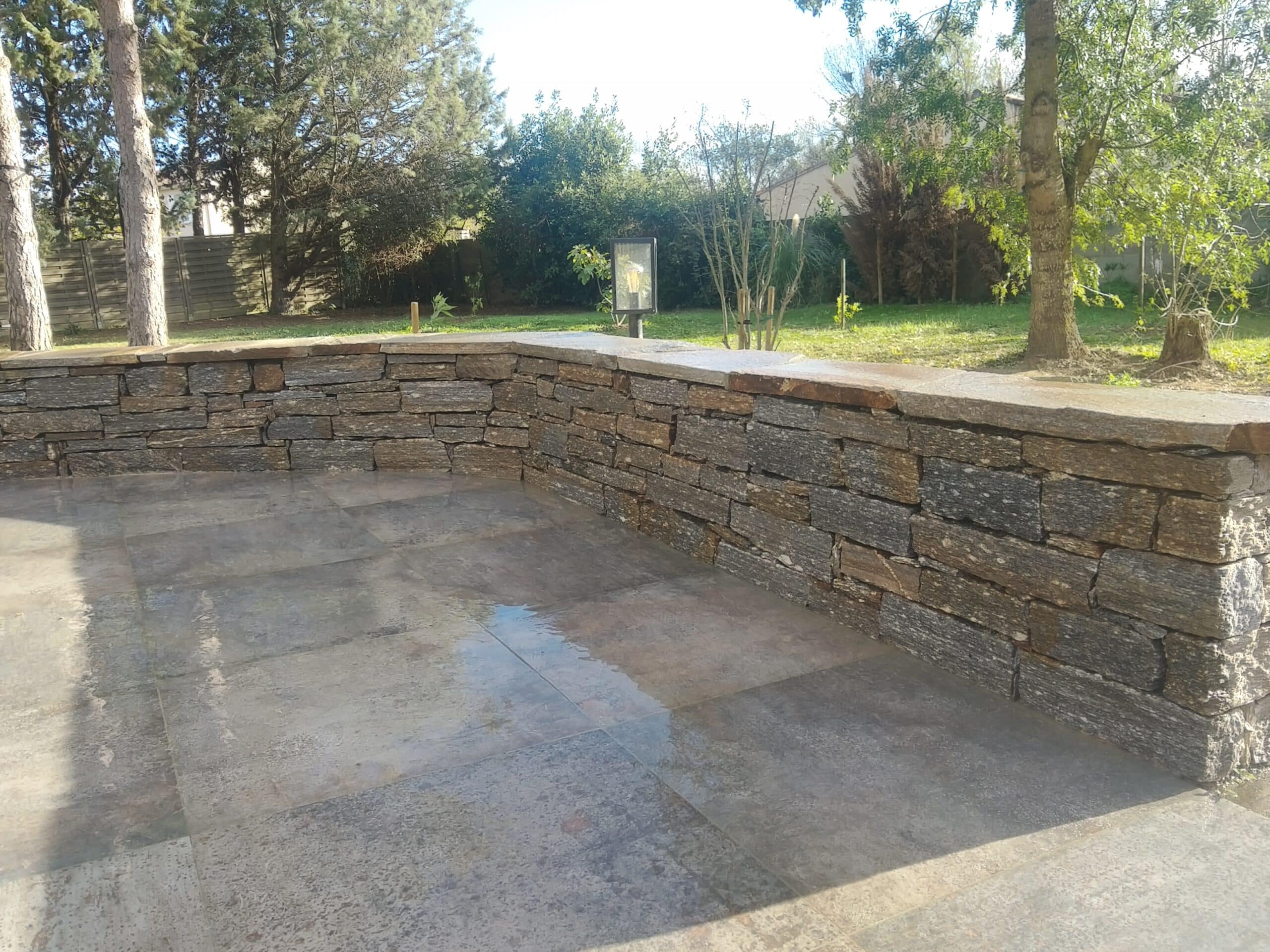 Terrasse en Pierre et Mur de Soutènement pour Jardin Terrasse moderne avec mur de soutènement en pierre sèche et dalles grises dans un jardin arboré ensoleillé.