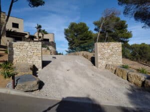 Entrée de villa avec allée en gravier et piliers en pierres sèches sous un ciel bleu dégagé.