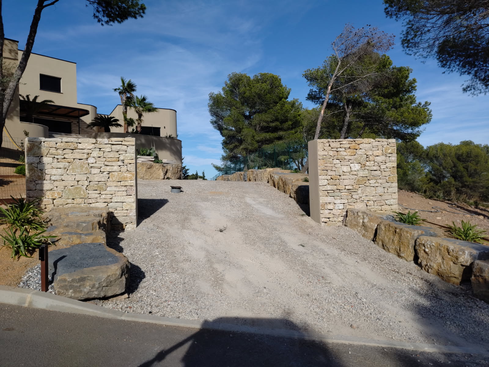 Entrée de villa avec allée en gravier et piliers en pierres sèches sous un ciel bleu dégagé.