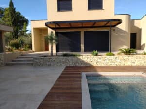 Villa moderne avec piscine, terrasse bois et muret en pierre sous pergola noire.