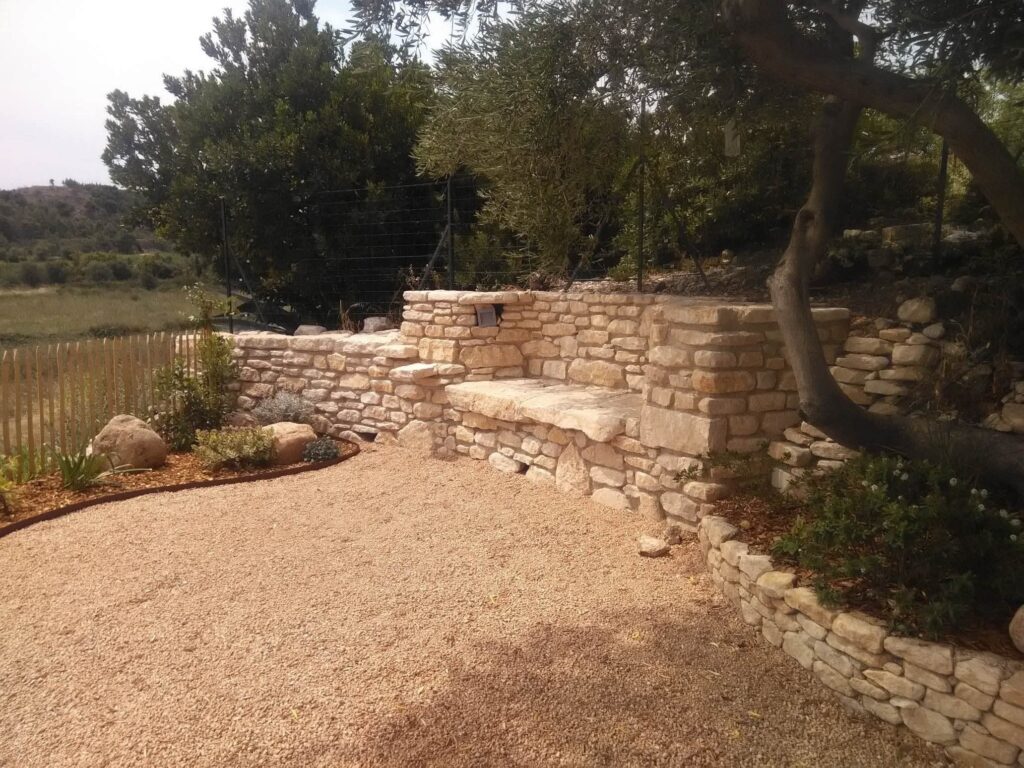 Aménagement jardin méditerranéen : muret et banc en pierre Banc et muret en pierre sèche dans un jardin paysager avec gravier et végétation méditerranéenne.