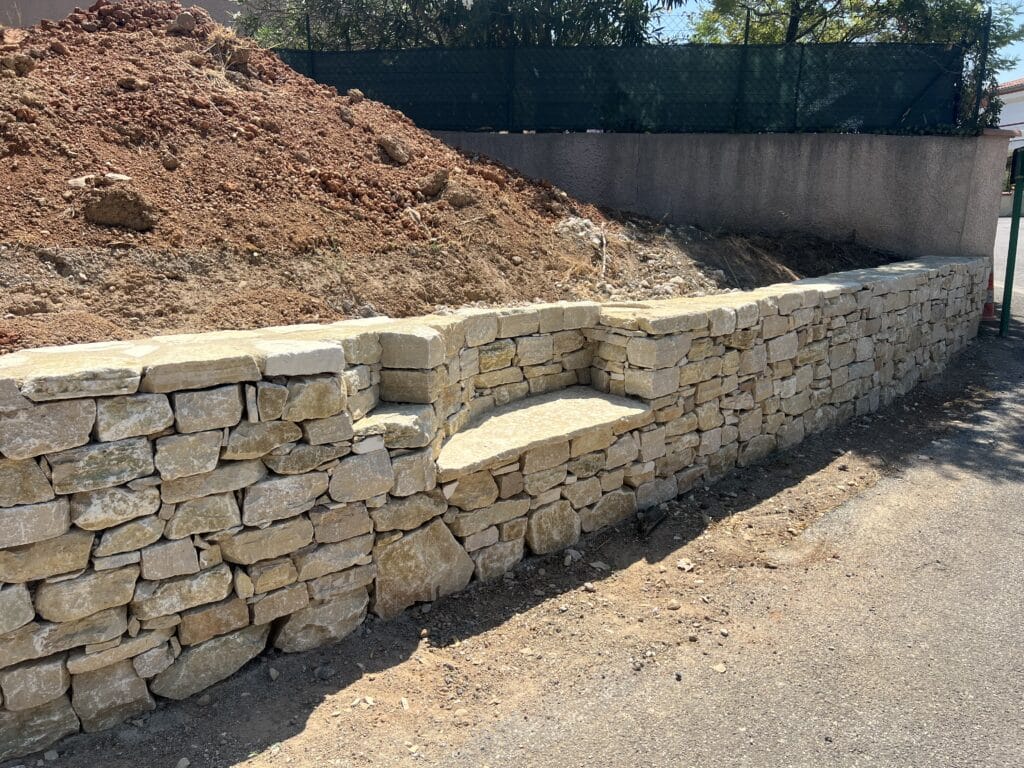 Mur en Pierre Sèche avec Banc : Aménagement de Jardin Mur en pierre sèche avec banc intégré devant un tas de terre rouge et une clôture verte.